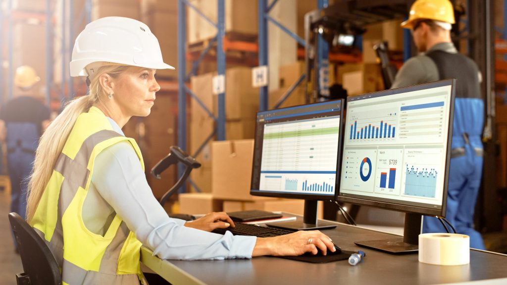 A logistics specialist wearing a hard hat and high-visibility vest working on a computer dashboard to monitor supplier delivery performance metrics.