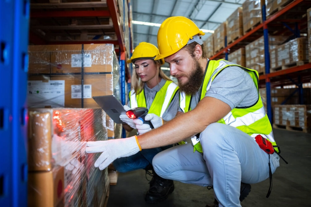 Two warehouse workers wearing hard hats and safety vests scanning barcodes on a pallet to prevent mismatched SKU warehouse transfers.