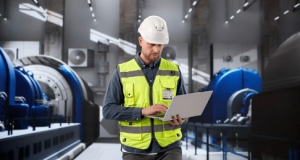 An industrial engineer using a laptop on a factory floor to audit maintenance logs and identify hidden work order cost capture failures.